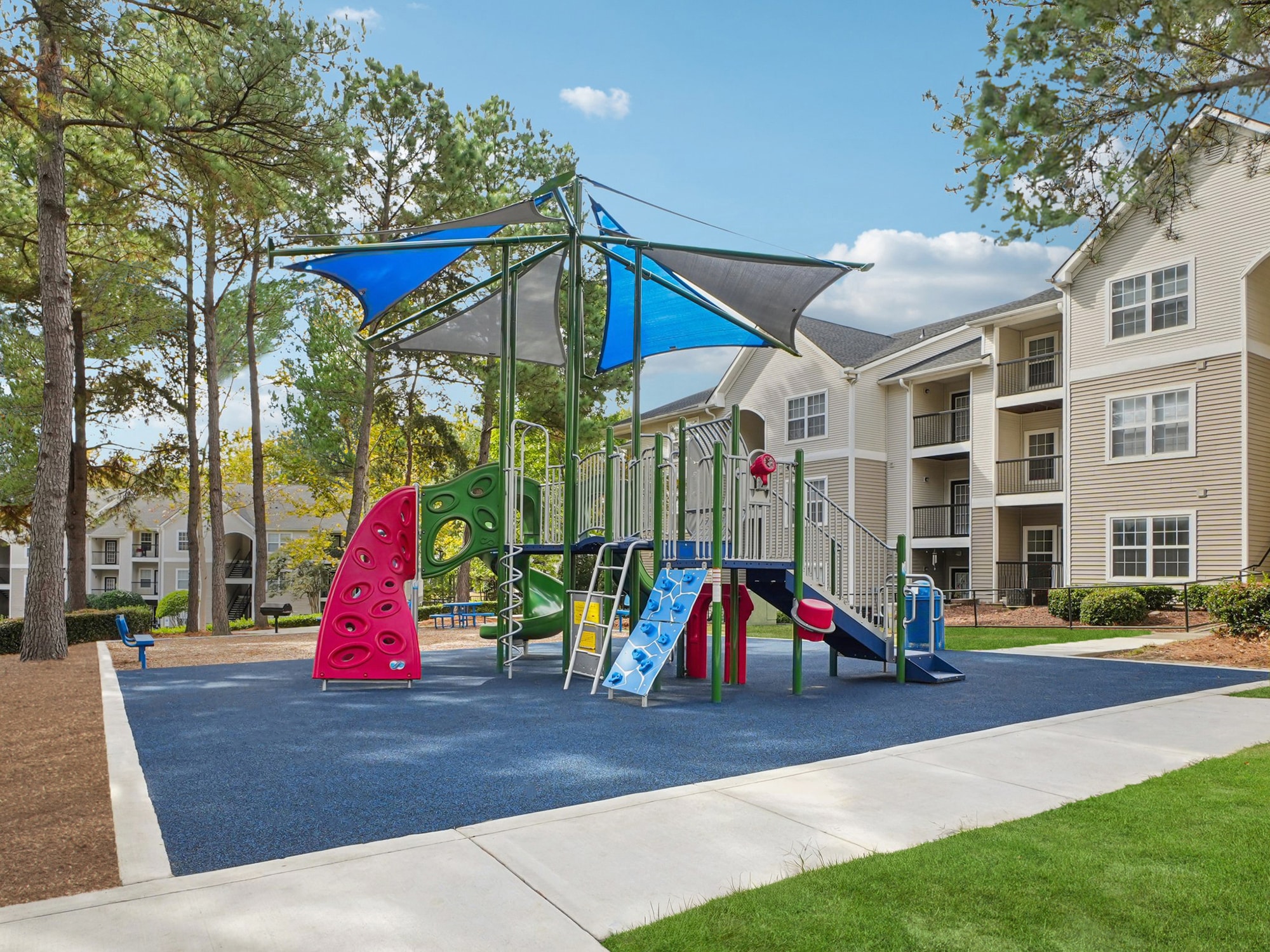 Colorful playground with slides, climbing wall, and shade canopy stands on blue rubber flooring near modern apartment buildings and trees under a clear sky.