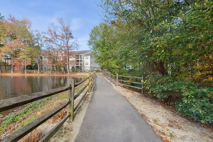 Paved walking path bordered by wooden fences runs alongside a pond, with trees and an apartment building in the background under a clear sky.