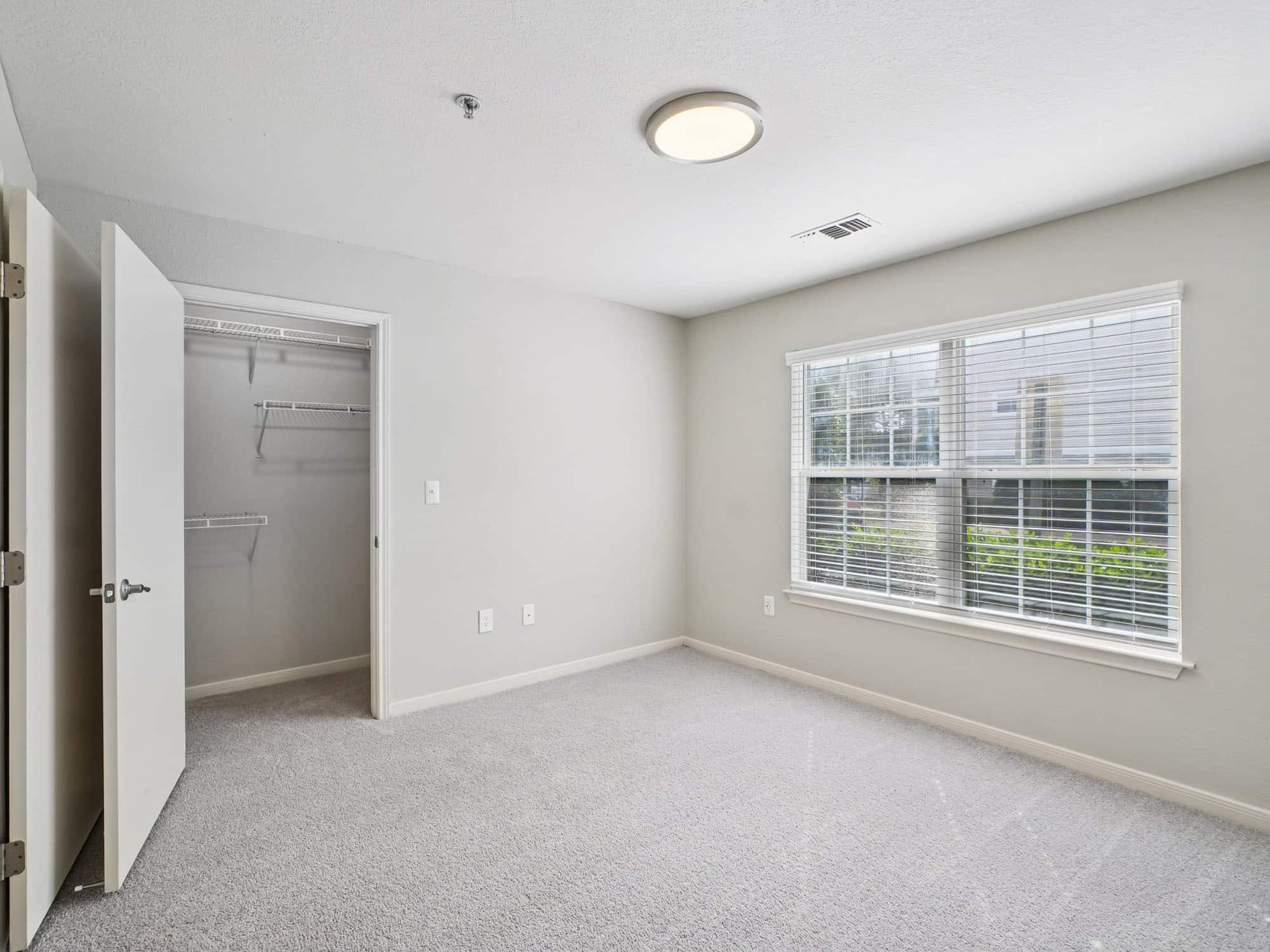 Empty bedroom with light gray carpet, open closet door, and a large window with blinds letting in natural light.
