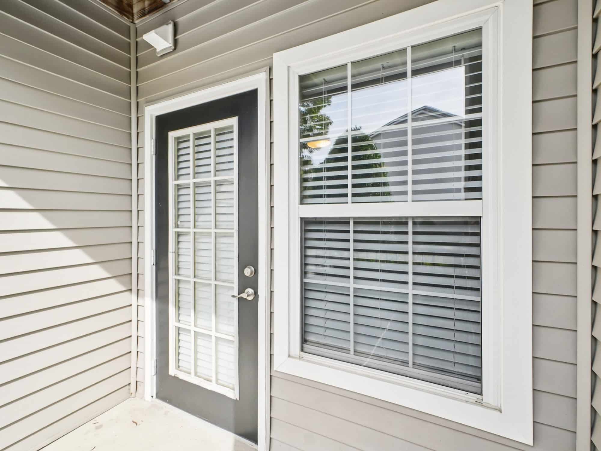 A glass-paneled door with white blinds next to a double-hung window with white trim, set in beige vinyl siding.