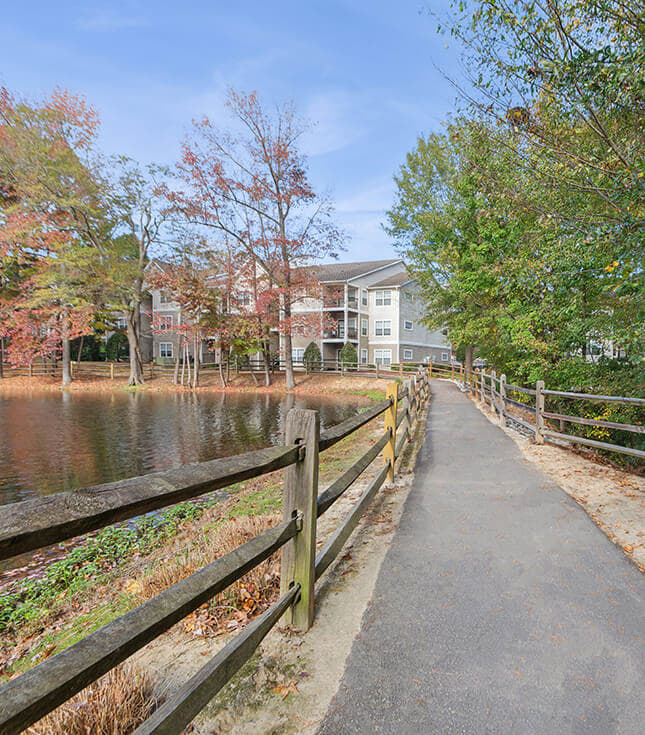 Paved walkway with wooden fence beside a pond, leading toward apartment buildings surrounded by trees with autumn foliage.