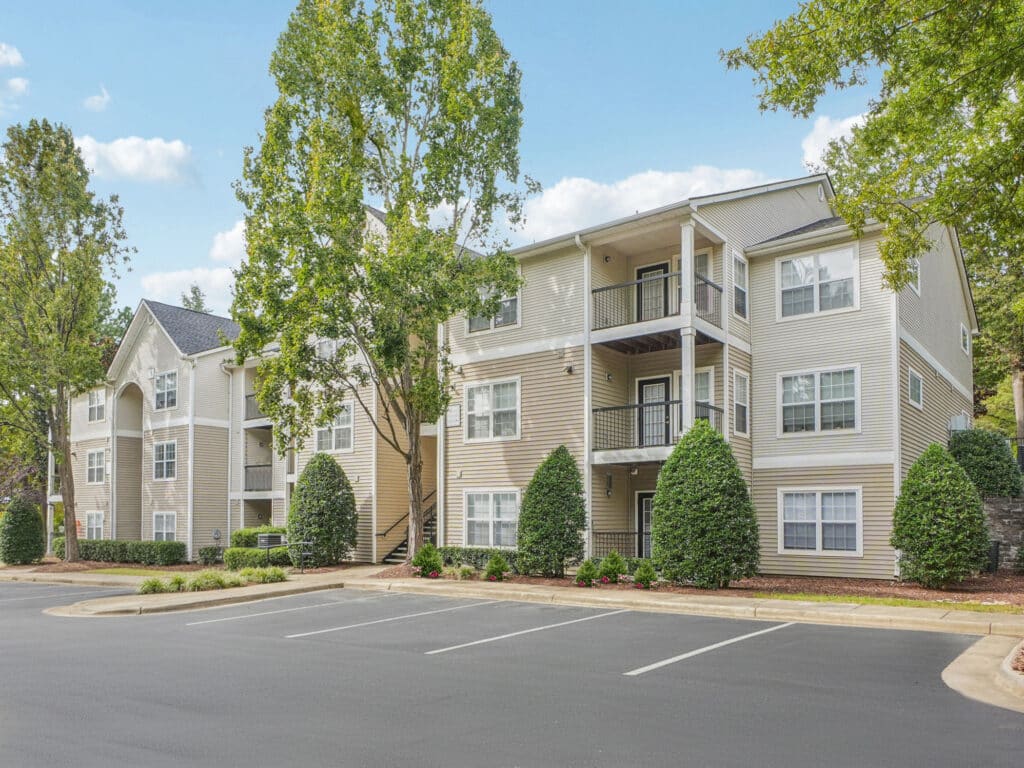 Three-story beige apartment building with balconies, surrounded by trees and shrubs, and adjacent to an empty asphalt parking lot under a blue sky.