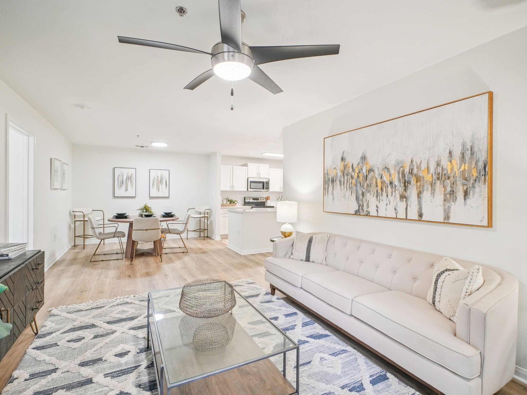 Modern open-concept living and dining area with neutral decor, a beige sofa, glass coffee table, abstract wall art, and a ceiling fan, leading into a white kitchen.