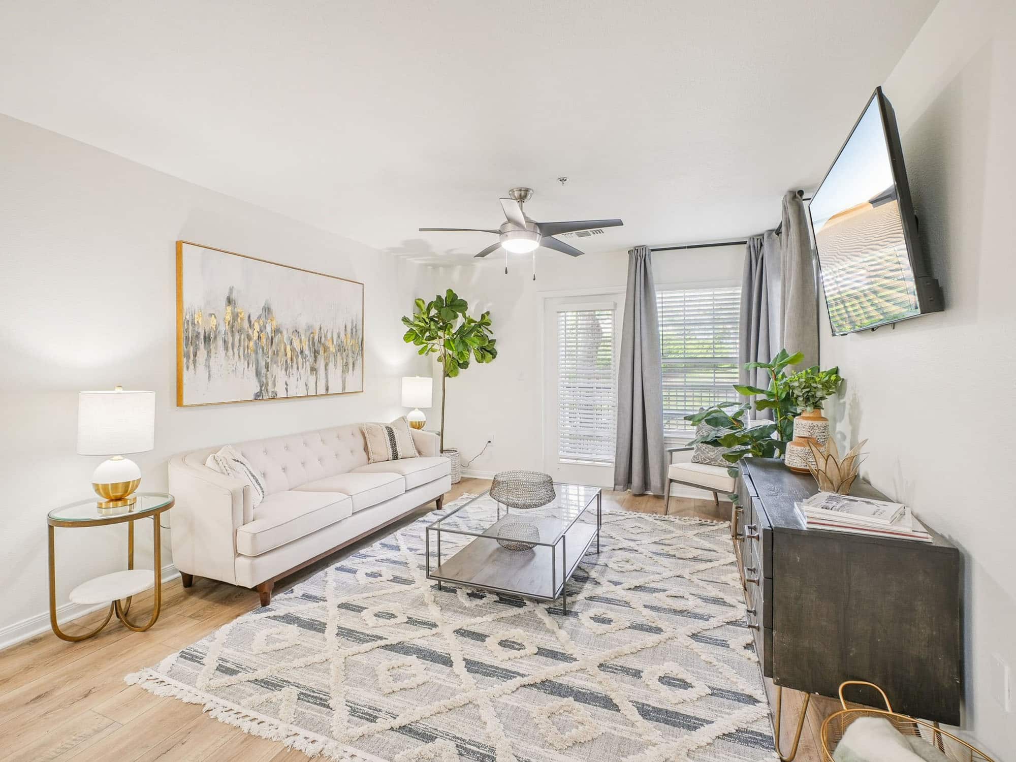 Bright living room with a white sofa, glass coffee table, wall-mounted TV, abstract artwork, large rug, floor plant, and natural light from windows with gray curtains.