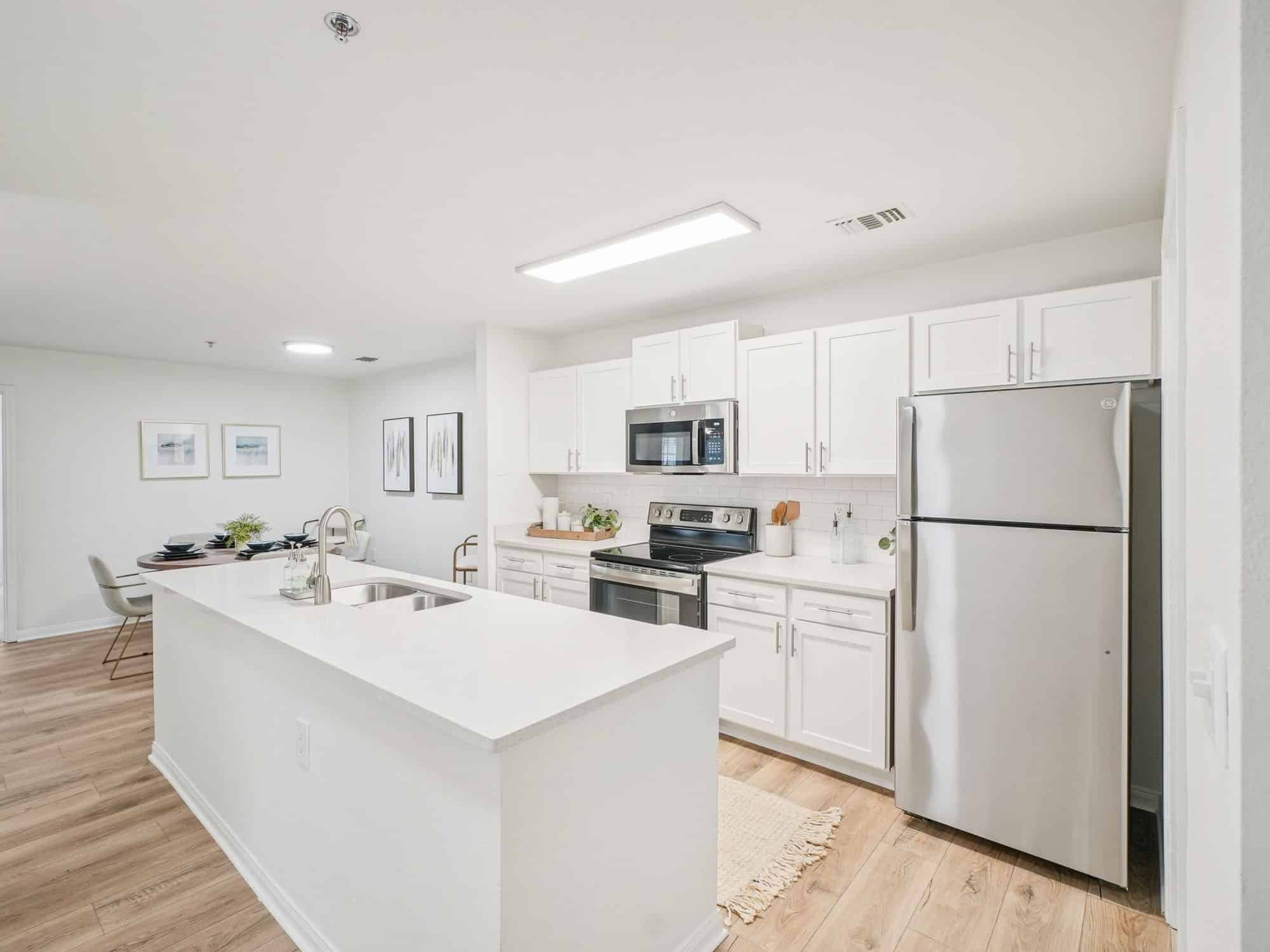 Modern kitchen with white cabinets, stainless steel appliances, an island with a sink, and light wood flooring; dining area visible in the background.