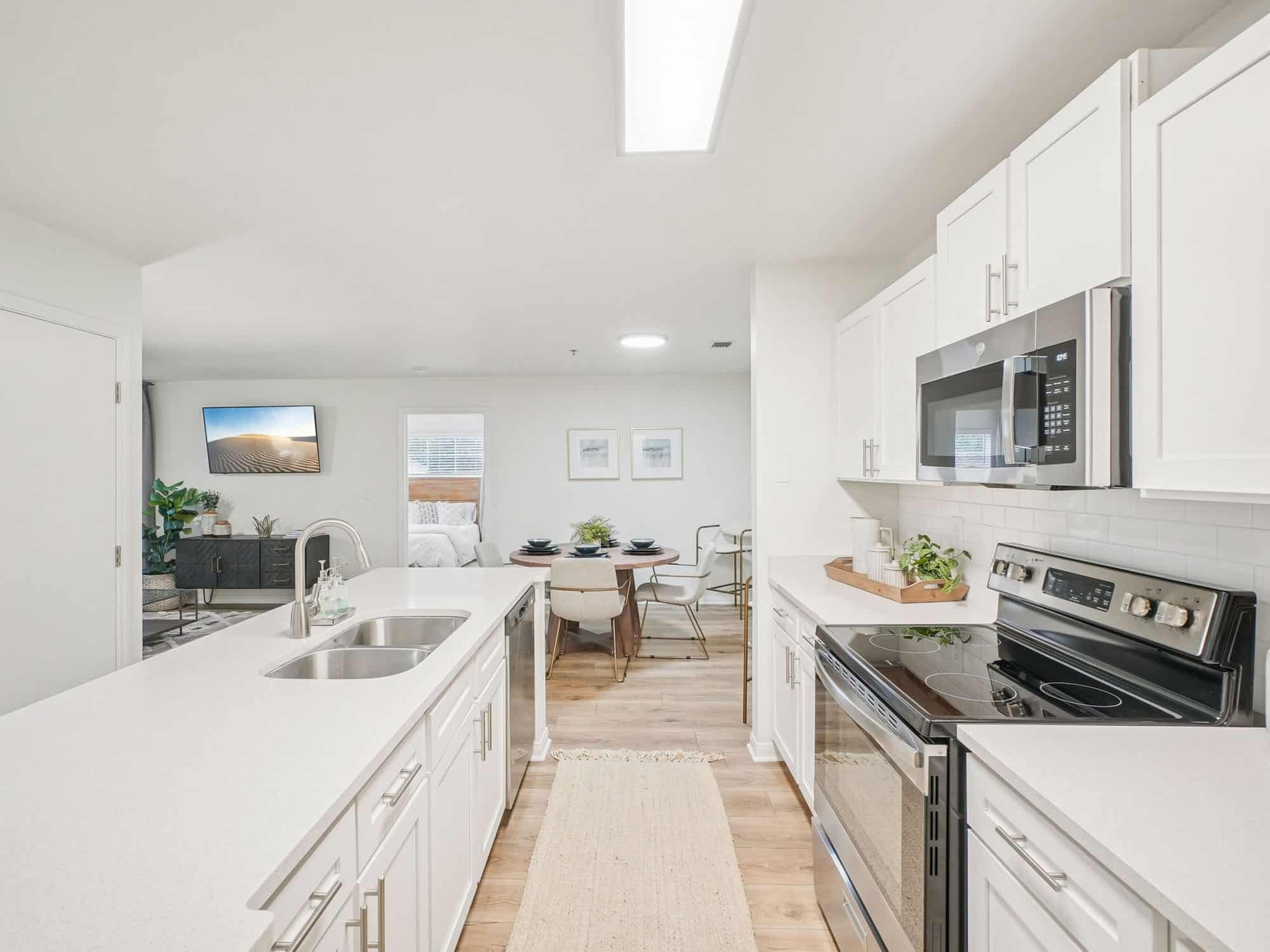Modern white kitchen with stainless steel appliances, a double sink island, and a view of the dining and living area in an open floor plan.