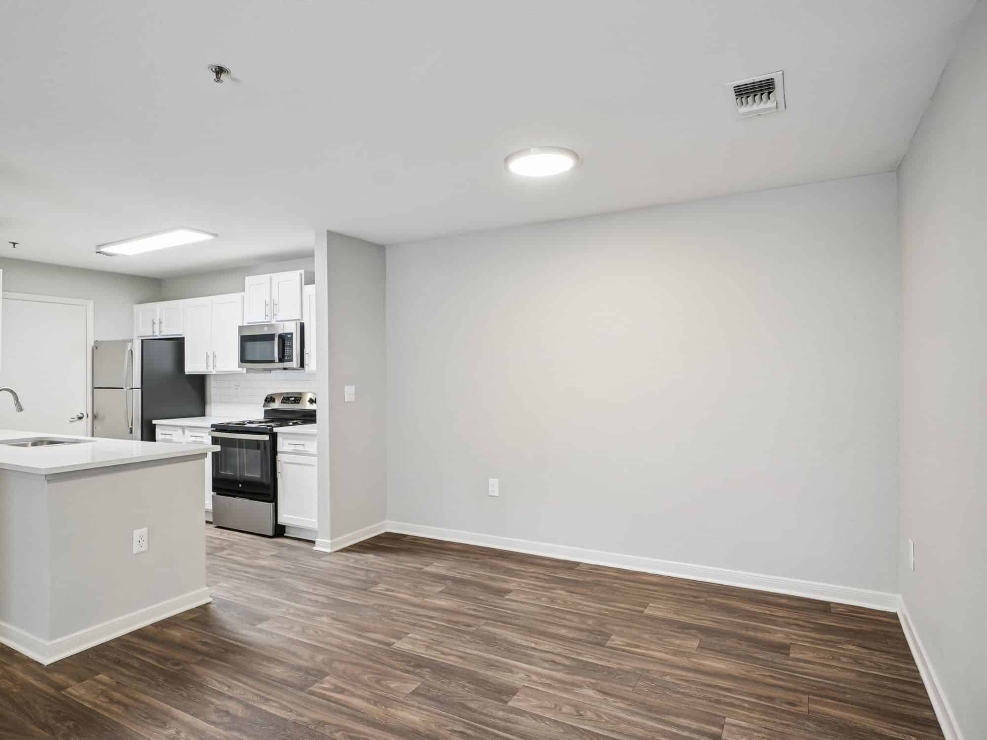 Modern apartment interior with an open kitchen featuring white cabinets and stainless steel appliances, empty living area, and wood-style flooring.