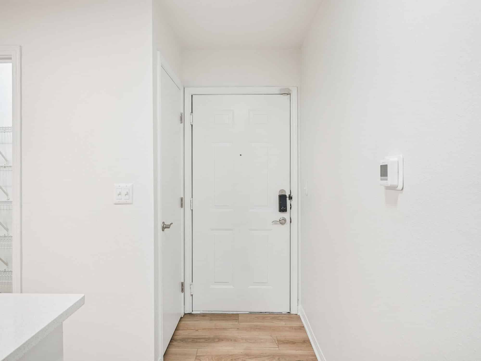 A white entry door with a silver handle and lock, flanked by a closet door on the left. The floor is wood, and the walls are white with a thermostat on the right wall.