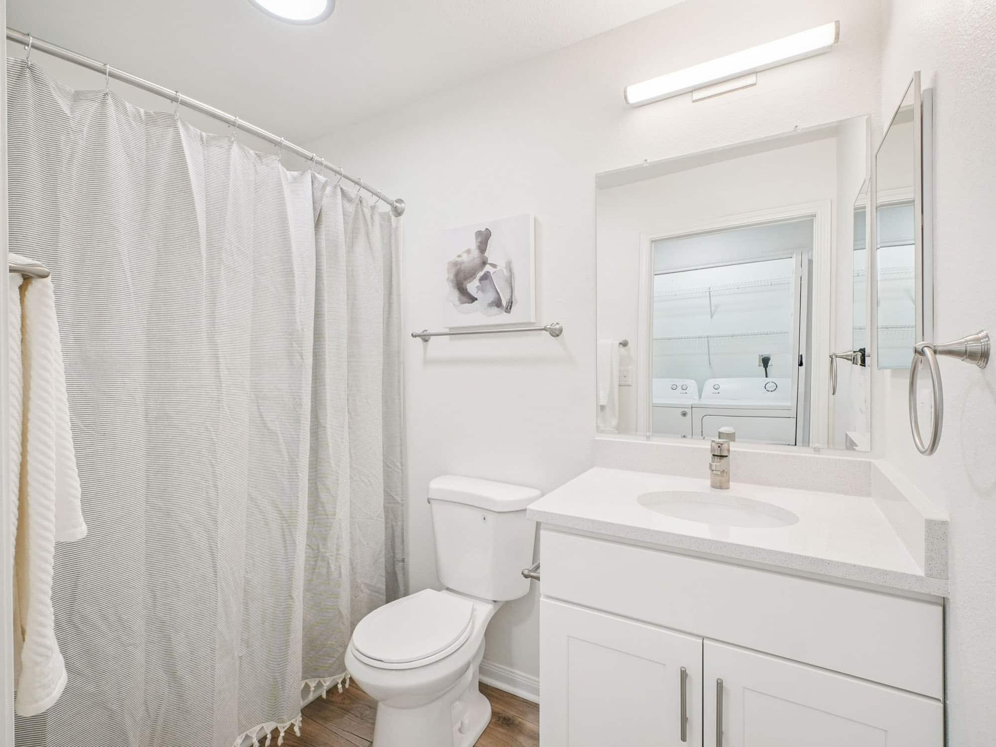 A clean, white bathroom with a shower curtain, toilet, sink with a mirror, wall art above the toilet, and a washing machine visible in the reflection.
