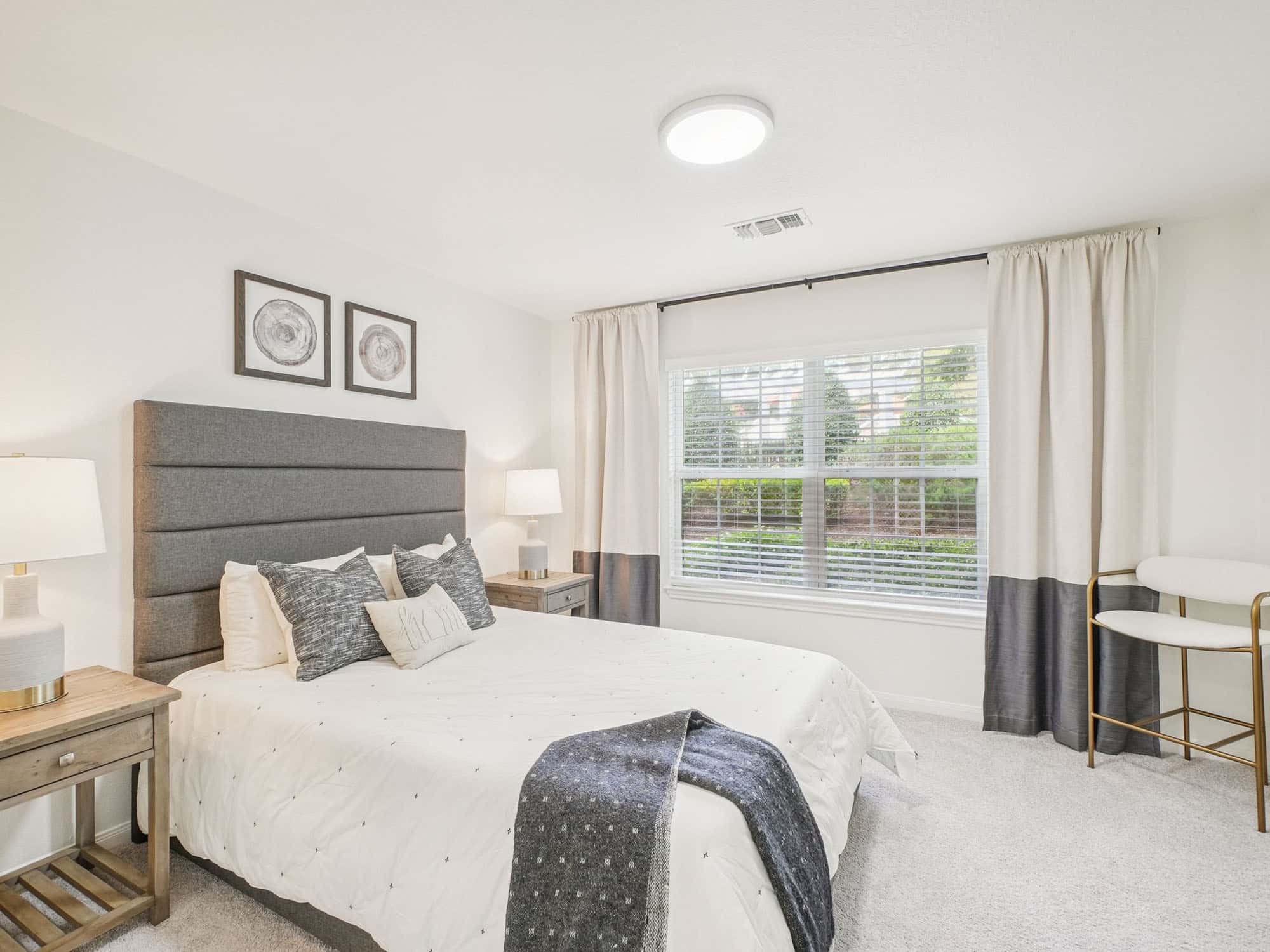 Bright bedroom with a large window, double bed with gray headboard, two bedside tables with lamps, and neutral decor. A chair and desk are positioned by the window.