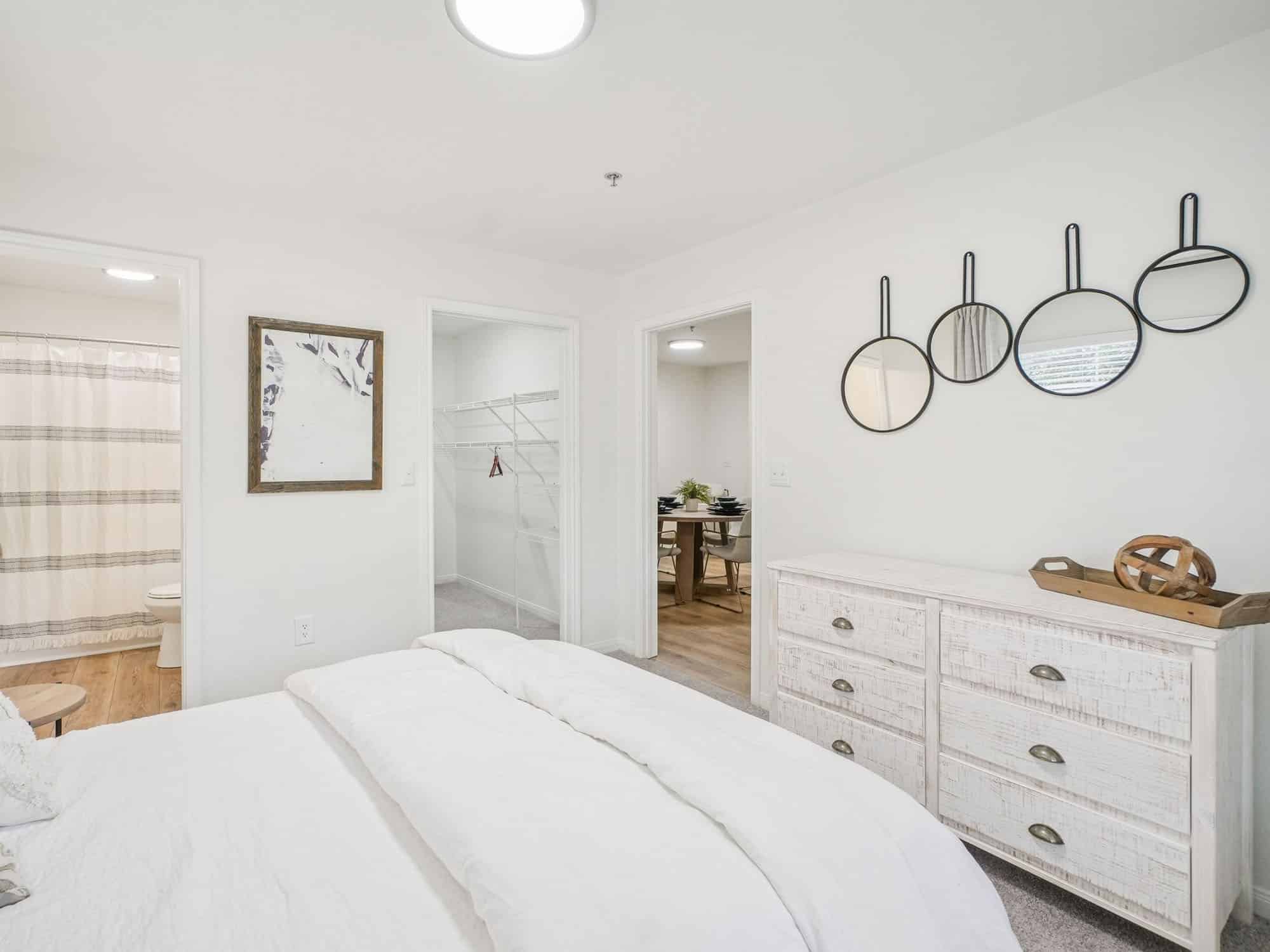 A bright bedroom with a white bed, a dresser, three round mirrors on the wall, and doorways to a bathroom, closet, and dining area.