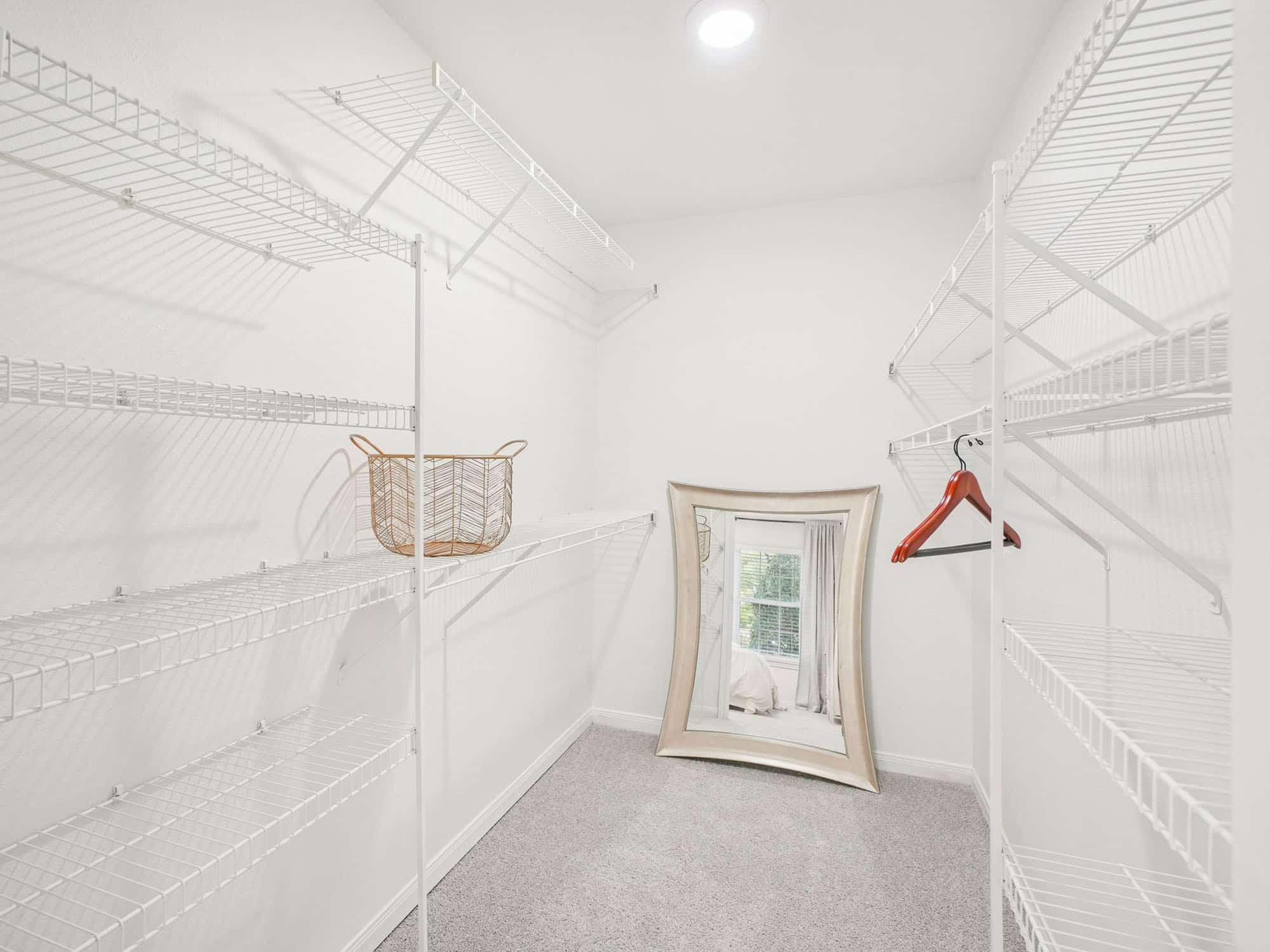 Empty walk-in closet with white wire shelves, a basket, a few wooden hangers, and a framed mirror resting against the far wall near a window.