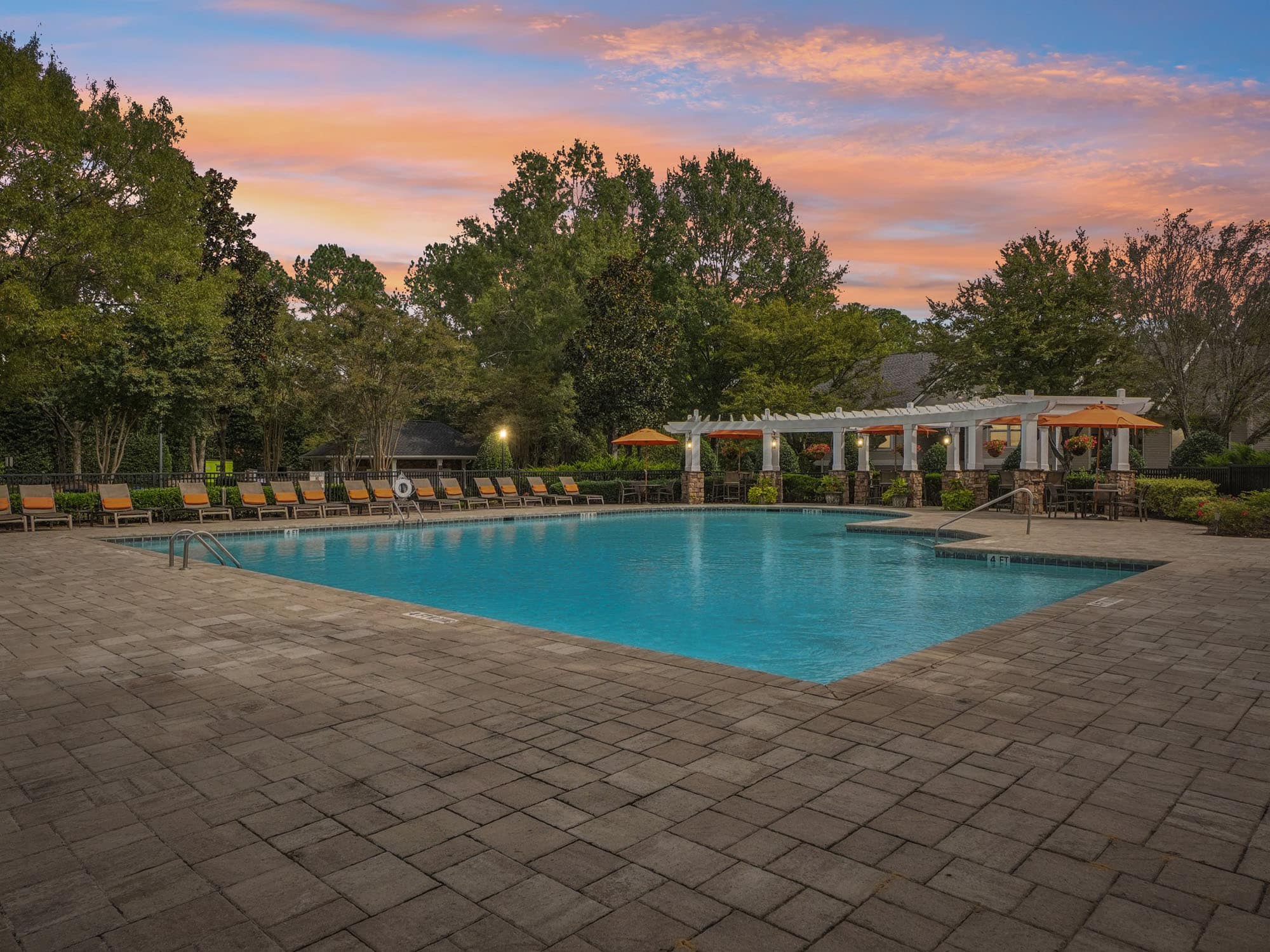 Rectangular outdoor swimming pool with lounge chairs, umbrellas, and a pergola area, surrounded by trees under a colorful sunset sky.