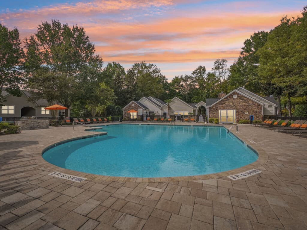 Outdoor swimming pool surrounded by lounge chairs and trees, with residential buildings in the background under a colorful sunset sky.