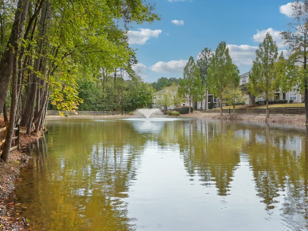 A pond with a water fountain in the center, surrounded by trees and residential buildings under a partly cloudy sky.