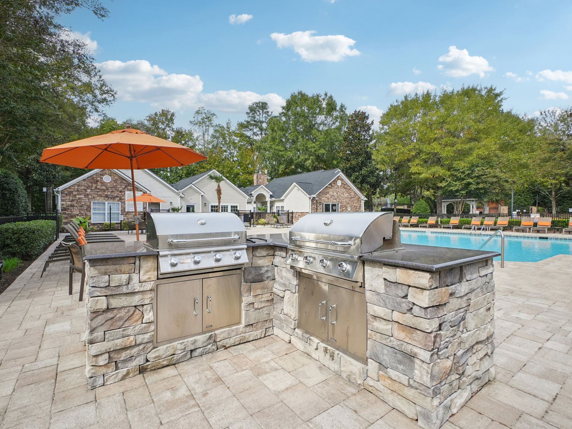 Two stainless steel grills built into a stone outdoor kitchen are positioned next to a swimming pool with lounge chairs and an orange patio umbrella in a landscaped residential area.