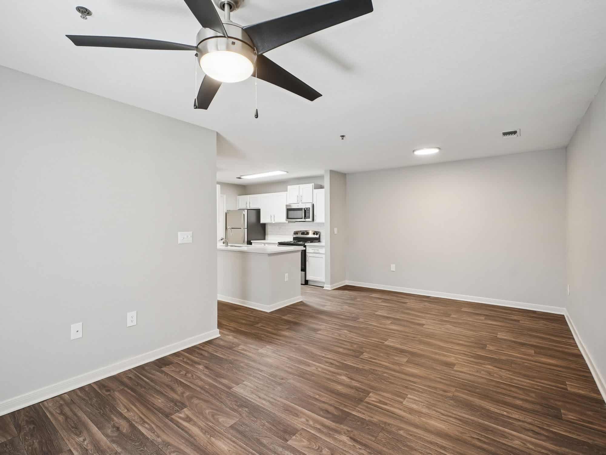 Empty apartment interior with wood flooring, neutral walls, a ceiling fan, and a view of a modern open kitchen with white cabinetry and stainless steel appliances.