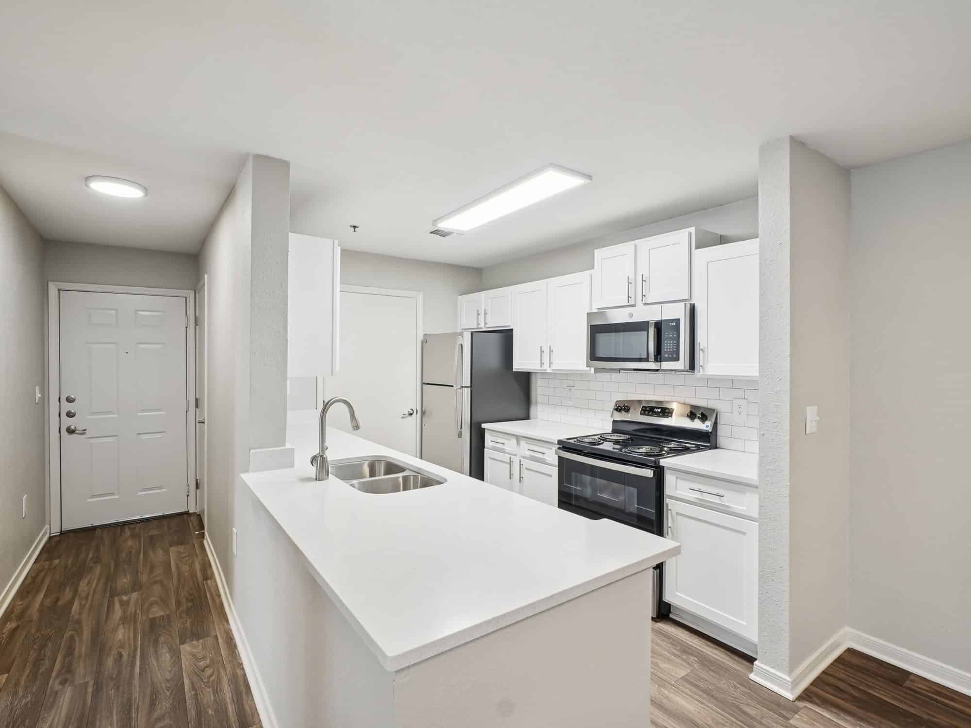 Modern kitchen with white cabinets, stainless steel appliances, island with sink, and wood-look flooring adjacent to entry door in a well-lit apartment.