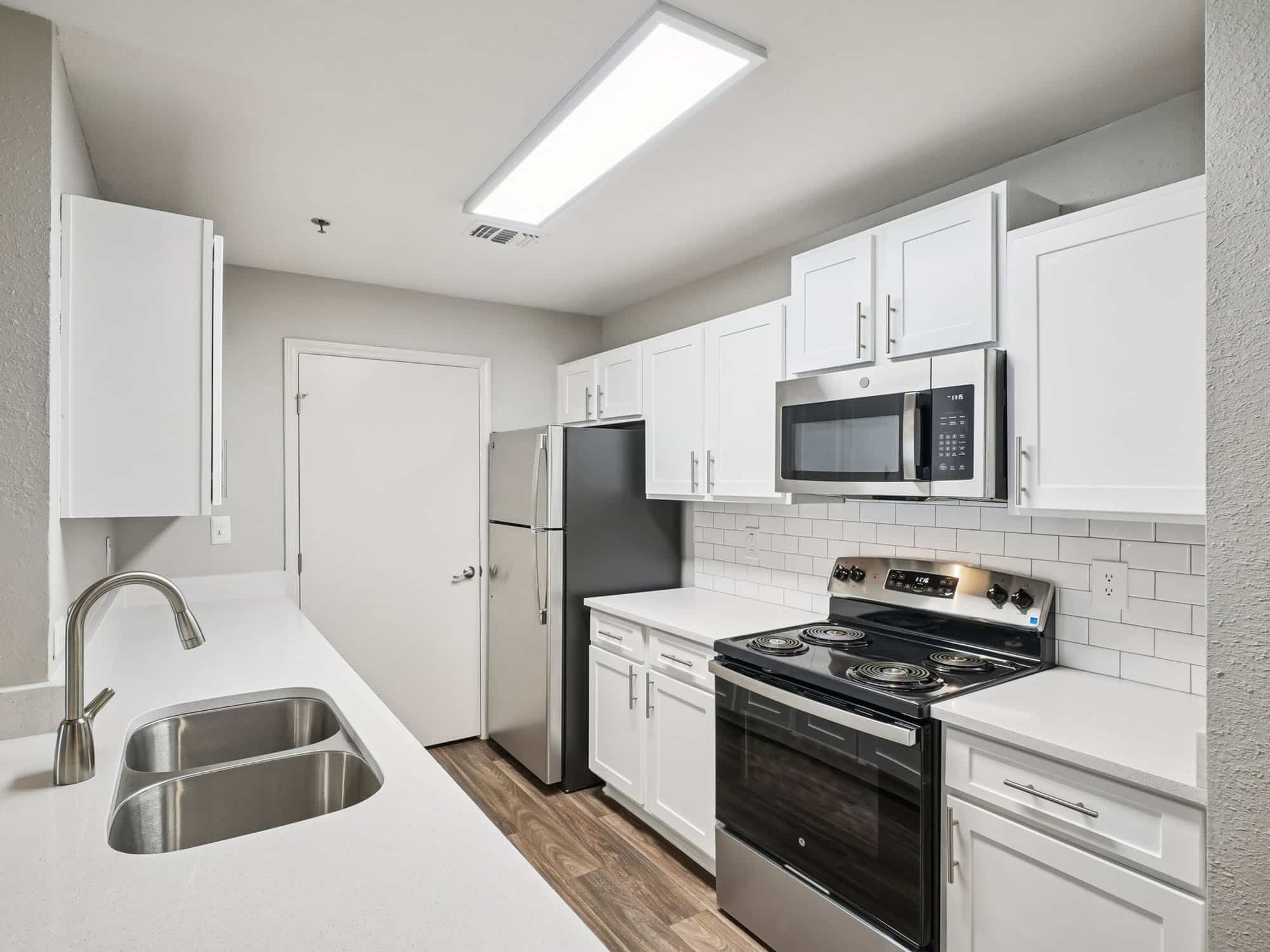 Modern kitchen with white cabinets, stainless steel appliances, electric stove, built-in microwave, double sink, and white countertops. Bright overhead lighting and neutral walls.